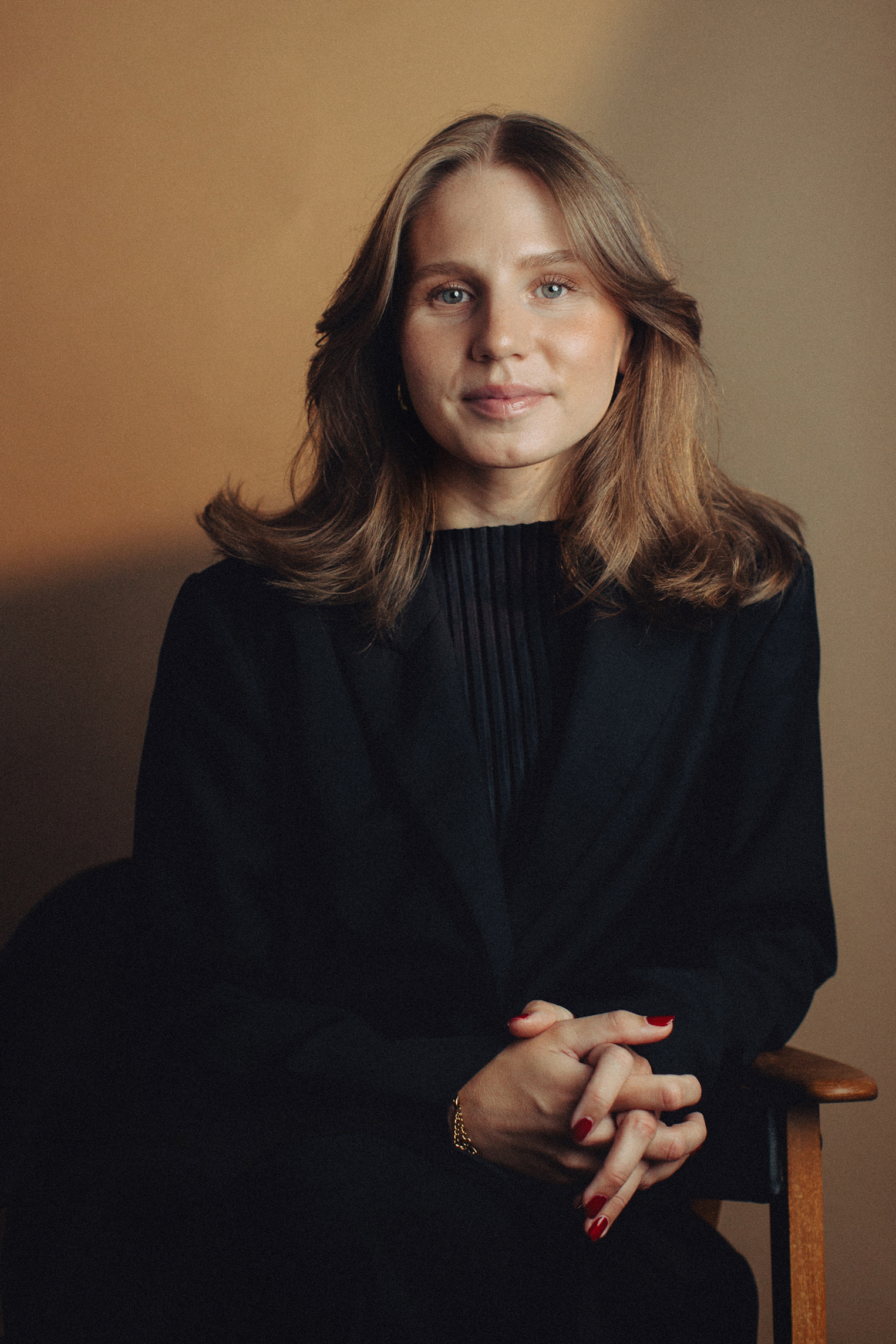 Evelina sitter på en stol och tittar in i kameran, hon har händerna knäppta och tittar lende in i kameran. Evelina är klädd i kostym.
