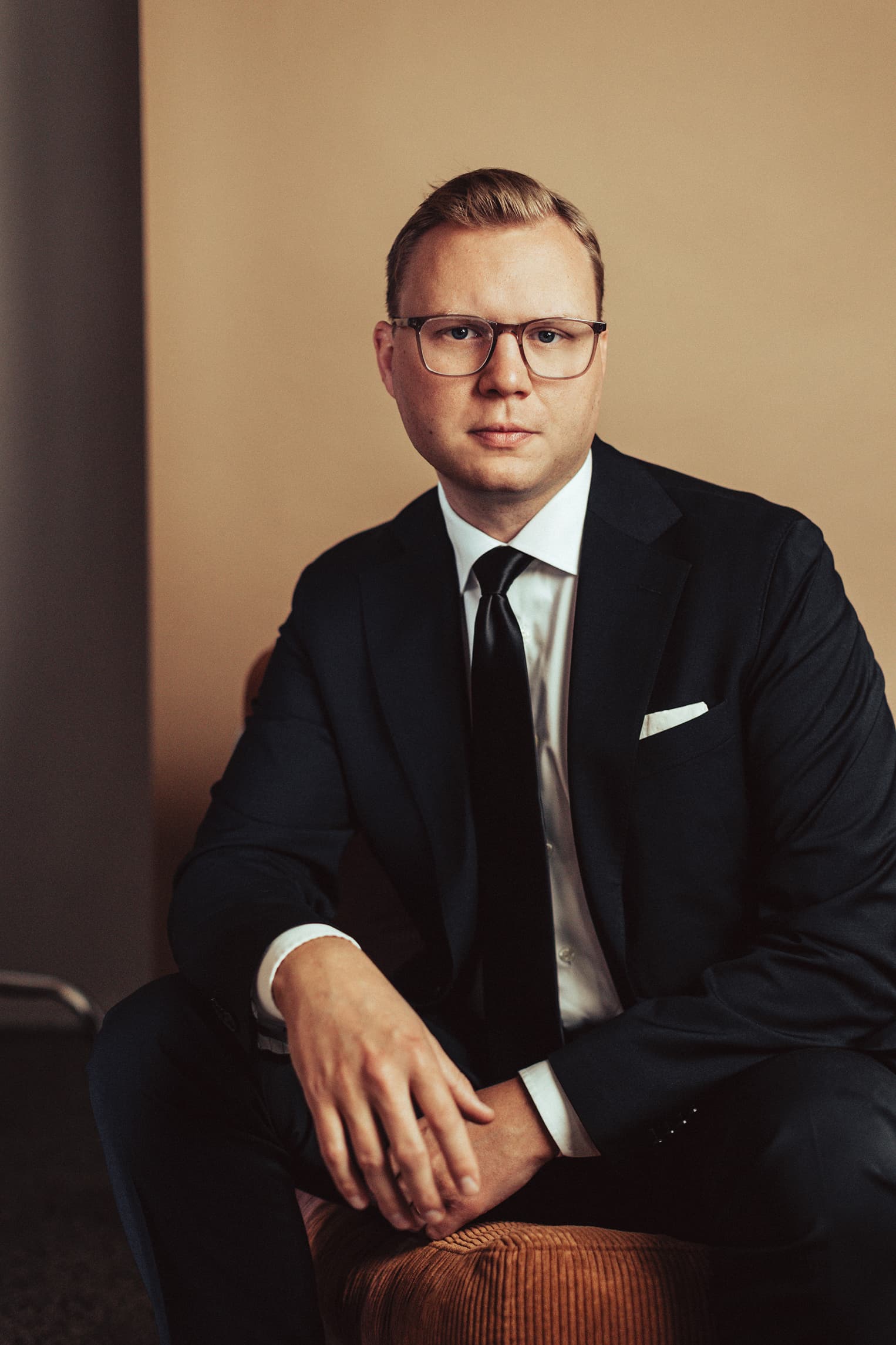 Sebastian sitter framåtlutad med händerna knäppta och tittar in i kameran. Sebastian är klädd i mörk kostym och glasögon.